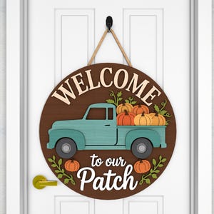 May include: A round wooden sign with the words "WELCOME to our Patch" in cream-colored letters. A teal pickup truck carries orange pumpkins. The sign hangs from a jute rope and is mounted on a white door.