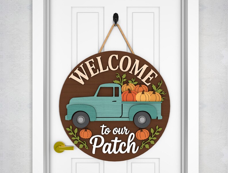 May include: A round wooden sign with the words "WELCOME to our Patch" in cream-colored letters. A teal pickup truck carries orange pumpkins. The sign hangs from a jute rope and is mounted on a white door.