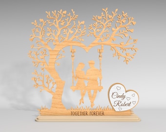 Puede incluir: Adorno decorativo de madera que representa un árbol con un dosel en forma de corazón y un columpio con dos figuras. La base dice "TOGETHER FOREVER" e incluye una placa en forma de corazón con nombres. La pieza es de madera clara.