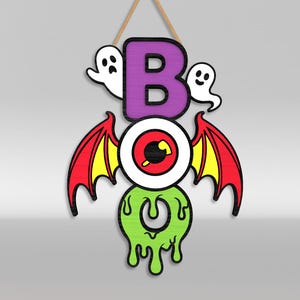 Op de afbeelding: Een hangend houten wandbord met de tekst BOO, bestaande uit een paarse letter B, een centraal oog met rode en gele vleermuisvleugels, en een druipende limoengroene letter O. Twee kleine witte spookjes bevinden zich aan weerszijden van de letter B.