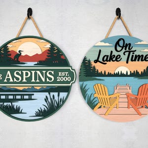 Pode incluir: Duas placas redondas de madeira com tema de lago com cabides de corda. Uma placa apresenta uma cena de lago com uma gar&ccedil;a e o texto "THE ASPINS EST. 2000". A outra placa diz "On Lake Time" com cadeiras Adirondack em um cais.
