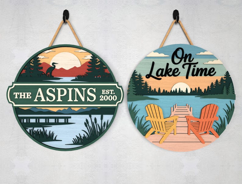 Pode incluir: Duas placas redondas de madeira com tema de lago com cabides de corda. Uma placa apresenta uma cena de lago com uma gar&ccedil;a e o texto "THE ASPINS EST. 2000". A outra placa diz "On Lake Time" com cadeiras Adirondack em um cais.