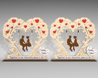 Puede incluir: Dos piezas decorativas de madera. Cada una presenta un diseño en forma de árbol con una corona en forma de corazón y detalles de corazón rojo. Siluetas de una pareja en un columpio están en el centro. El texto "Together is our favourite place to be" está en la base.
