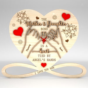 May include: Wooden heart-shaped sign with the text "THE Mother & Daughter BOND IS A knot TIED BY ANGEL'S HANDS I LOVE YOU MOM" and two hands holding a red knot. The names "Mommy" and "Anna" are written on the hands.