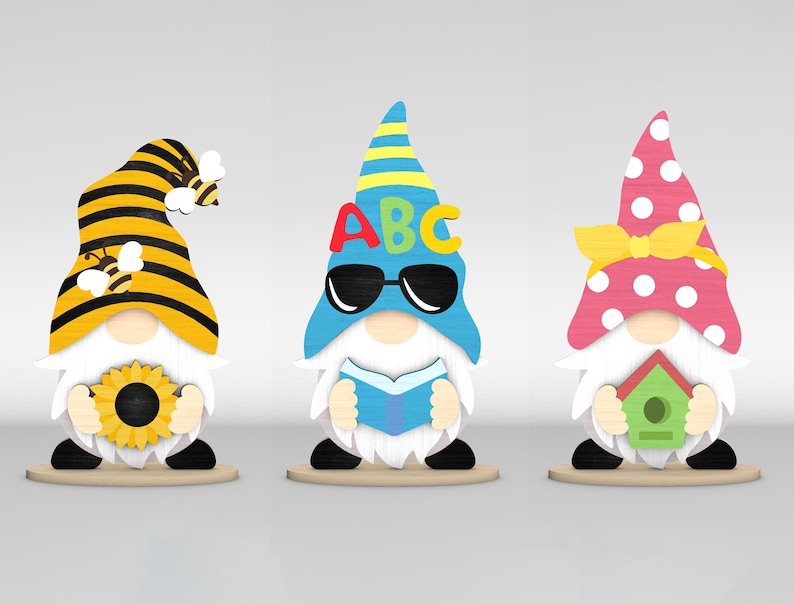 May include: Three wooden gnome figurines. One gnome wears a yellow and black striped hat with a bee and holds a sunflower. Another gnome has a blue hat with the letters ABC and sunglasses, holding a book. The third gnome wears a pink polka-dot hat and holds a birdhouse.