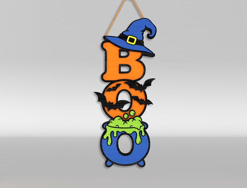Op de afbeelding: Een verticaal hangend wandbord met de tekst BOO. De letter B is oranje met een blauwe heksenhoed. De letter O is oranje met zwarte vleermuissilhouetten. De laatste O is een blauwe ketel met groene borrelende vloeistof, opgehangen aan een touw.