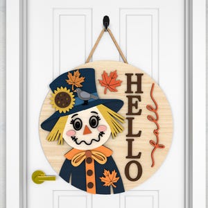 May include: A round wooden door sign featuring a scarecrow design. The scarecrow has a blue hat with a sunflower and bird, and a straw-like face. The word "HELLO" is written on the sign, with fall leaves.
