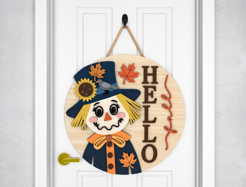 May include: A round wooden door sign featuring a scarecrow design. The scarecrow has a blue hat with a sunflower and bird, and a straw-like face. The word "HELLO" is written on the sign, with fall leaves.