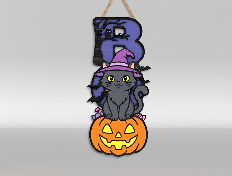 Op de afbeelding: Een hangend houten bord met de letter B in paars met een zwart boomsilhouet en een kleine kat. Een grijze kat met een paarse heksenhoed zit op een oranje pompoen met gele ogen en mond, omringd door kleine snoepjes.