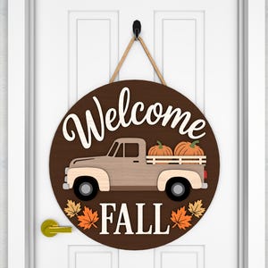 May include: A round wooden door sign with a brown background and the words "Welcome Fall" in white. The sign features a light brown truck carrying two pumpkins. The sign is decorated with orange and yellow fall leaves.