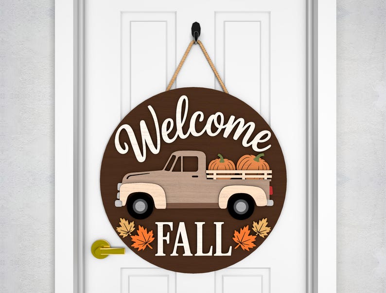 May include: A round wooden door sign with a brown background and the words "Welcome Fall" in white. The sign features a light brown truck carrying two pumpkins. The sign is decorated with orange and yellow fall leaves.