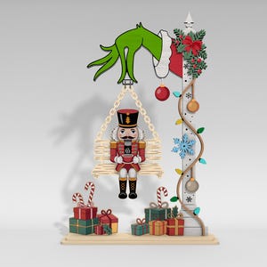 Può includere: Decorazione natalizia in legno raffigurante uno schiaccianoci su un'altalena, tenuto da una mano verde. Un palo decorato con ornamenti e regali completa la scena festosa. Lo schiaccianoci tiene in mano una tazza.