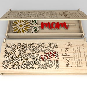 Puede incluir: Una caja de madera con un diseño floral cortado con láser. La caja está abierta, revelando una flor y la palabra "mamá" en rojo. La tapa tiene la palabra "madre" y una definición. La caja es de madera clara.