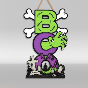 Op de afbeelding: Een verticaal hangbord met het woord BOO in grote letters. De B is limoengroen met zwarte stiksels en gekruiste botten. De O is paars met een groene zombiehand die eruit steekt. De laatste O is paars met een kerkhofsc&egrave;ne aan de onderkant.