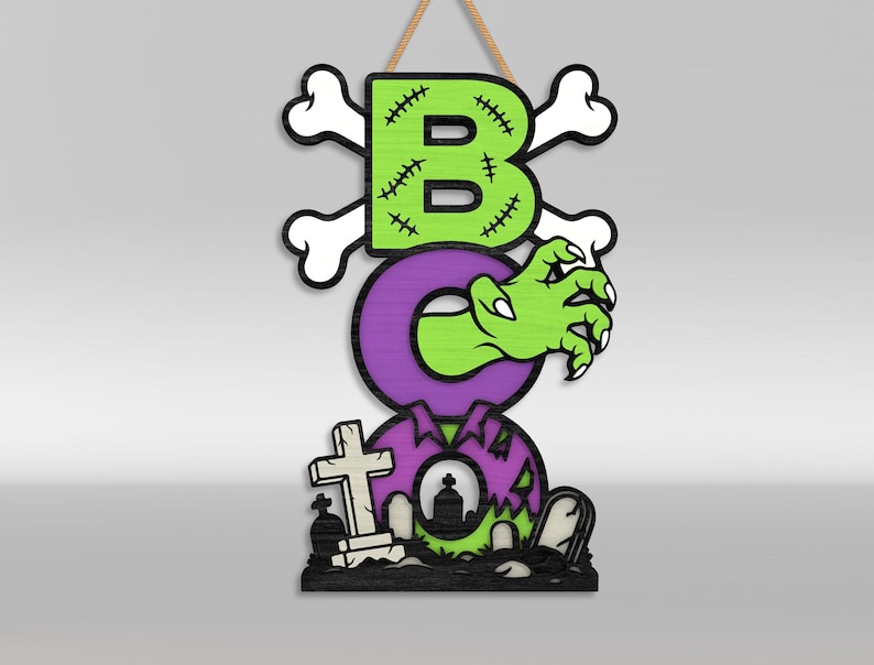 Op de afbeelding: Een verticaal hangbord met het woord BOO in grote letters. De B is limoengroen met zwarte stiksels en gekruiste botten. De O is paars met een groene zombiehand die eruit steekt. De laatste O is paars met een kerkhofsc&egrave;ne aan de onderkant.