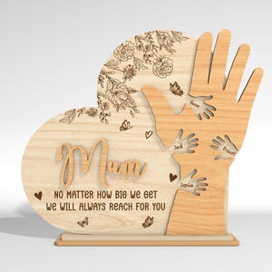 May include: Wooden heart-shaped plaque with the word "Mum" and a message that reads "No matter how big we get we will always reach for you". The plaque features a design of hands reaching up towards the word "Mum" with names inscribed on each hand.