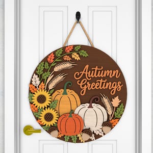 May include: A round wooden door sign with the words "Autumn Greetings" in orange script. The sign features sunflowers, pumpkins, leaves, and wheat stalks in fall colors. A rope is attached for hanging.