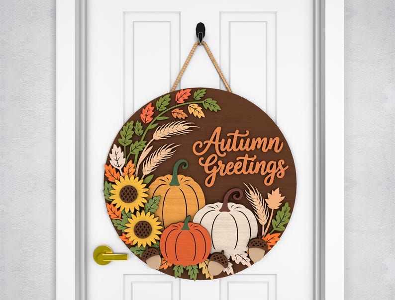 May include: A round wooden door sign with the words "Autumn Greetings" in orange script. The sign features sunflowers, pumpkins, leaves, and wheat stalks in fall colors. A rope is attached for hanging.