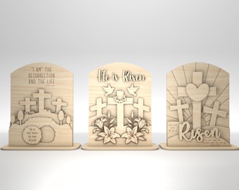 May include: Three wooden signs with a Christian Easter theme. The first sign has the text "I AM THE RESURRECTION AND THE LIFE" with three crosses and a tomb. The second sign has the text "He is Risen" with three crosses and lilies. The third sign has the text "Risen" with three crosses and a heart.