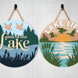 Pode incluir: Duas placas redondas de madeira com tema de lago. Uma diz "gone to the lake" com p&aacute;ssaros e uma cena de lago. A outra diz "lake life" com um p&ocirc;r do sol, p&aacute;ssaros e juncos. Cada placa tem um cabide de corda.