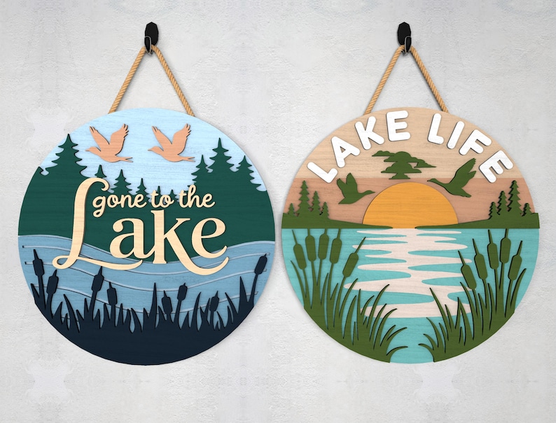 Pode incluir: Duas placas redondas de madeira com tema de lago. Uma diz "gone to the lake" com p&aacute;ssaros e uma cena de lago. A outra diz "lake life" com um p&ocirc;r do sol, p&aacute;ssaros e juncos. Cada placa tem um cabide de corda.