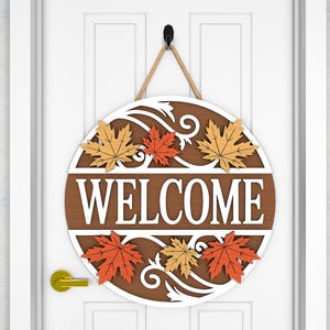May include: A round wooden welcome sign with a brown background and white lettering. The word "WELCOME" is prominently displayed. The sign is decorated with orange and yellow maple leaves and a white decorative border. It hangs from a rope.