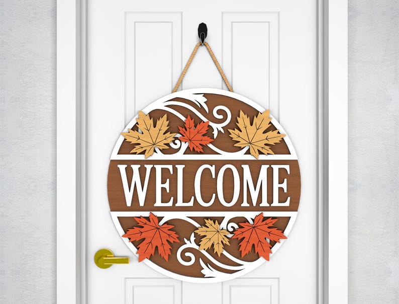 May include: A round wooden welcome sign with a brown background and white lettering. The word "WELCOME" is prominently displayed. The sign is decorated with orange and yellow maple leaves and a white decorative border. It hangs from a rope.