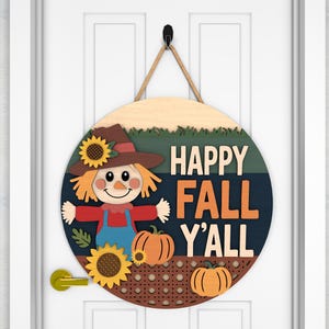 May include: Round wooden fall-themed door sign with a scarecrow, sunflowers, and pumpkins. The sign reads "HAPPY FALL Y'ALL" in orange and white lettering. The sign is hanging on a white door with a black hook and rope.