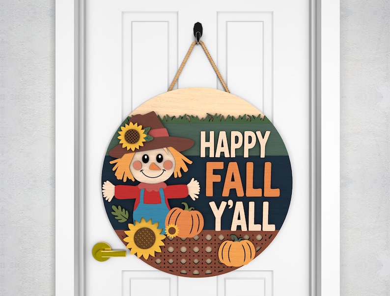 May include: Round wooden fall-themed door sign with a scarecrow, sunflowers, and pumpkins. The sign reads "HAPPY FALL Y'ALL" in orange and white lettering. The sign is hanging on a white door with a black hook and rope.