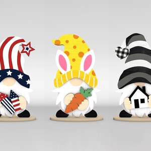 May include: Three wooden gnome figurines. One wears a red, white, and blue hat with a star and holds fireworks. Another has a yellow hat with bunny ears and holds a carrot. The third gnome has a black and white hat and holds a house.