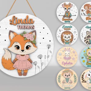 Puede incluir: Un letrero redondo de madera con un zorro de dibujos animados con un vestido rosa y un adorno floral. El letrero tiene el nombre "Linda Thomas" en letras naranjas. Letreros redondos más pequeños muestran varios personajes de animales con nombres.