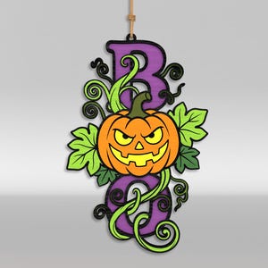 Op de afbeelding: Een hangend houten bord met het woord BOO in paarse blokletters en een oranje jack-o'-lantern pompoen in het midden. Het ontwerp bevat felgroene bladeren en donkere kronkelende ranken, opgehangen aan een dun beige touw.