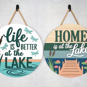 Pode incluir: Duas placas redondas de madeira com cabos de suspens&atilde;o. Uma placa diz "Life is Better at the Lake" com lib&eacute;lulas e juncos. A outra placa diz "Home is at the Lake" com um cais e nen&uacute;fares.