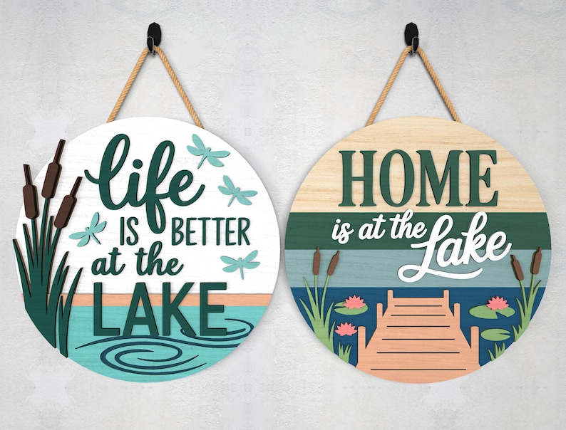 Pode incluir: Duas placas redondas de madeira com cabos de suspens&atilde;o. Uma placa diz "Life is Better at the Lake" com lib&eacute;lulas e juncos. A outra placa diz "Home is at the Lake" com um cais e nen&uacute;fares.