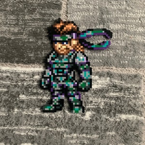 Op de afbeelding: Een pixel art-personage van een videogamepersonage dat een grijs en blauw pak draagt met een paars en blauw hoofdband. Het personage staat met zijn armen langs zijn zijde.