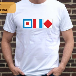 May include: A white short-sleeved t-shirt featuring a graphic print of three nautical signal flags arranged horizontally. The first flag is blue with a white square and red center, the second is vertical stripes of red, white, and blue, and the third is a red diamond on a white background.
