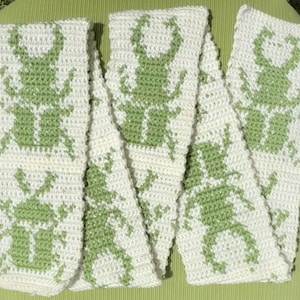 May include: A cream-colored, crocheted scarf featuring a repeating pattern of green insect figures. The scarf is folded and arranged on a green surface. The insect design is a simple, stylized silhouette.