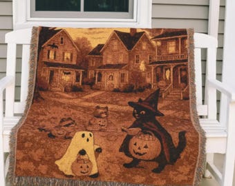 May include: A Halloween-themed throw blanket featuring a sepia-toned illustration of a neighborhood scene with trick-or-treaters. The blanket depicts a black cat in a witch hat, a ghost, and other characters holding jack-o'-lanterns. The blanket has a fringed edge.