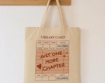 May include: Beige canvas tote bag with a library card design. The design includes the words "LIBRARY CARD", "JUST ONE MORE CHAPTER", and a heart. The bag has a rectangular shape and a handle for carrying.