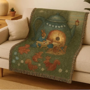 May include: A green throw blanket featuring a whimsical illustration of rabbits, squirrels, and a teapot. The blanket has a fringed edge and is draped over a beige sofa. The artwork has a vintage, storybook aesthetic.