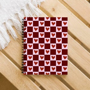 May include: A spiral-bound notebook with a red and pink checkered pattern, each square featuring a red heart. The notebook lies on a wooden surface, with a white fringed cloth in the background. The design is perfect for Valentine's Day.