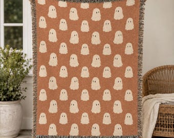 May include: A woven throw blanket featuring a repeating pattern of small, off-white ghost figures against a solid burnt orange background. The rectangular blanket has a textured knit appearance and is finished with a decorative fringe along the top and bottom edges.