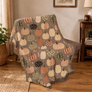 Vintage-inspired pumpkin woven blanket with decorative patchwork pumpkins surrounded by vibrant fall florals and foliage. The warm earthy palette and fringed edges create a cozy seasonal throw for chilly evenings, Halloween decorating, or Thanksgiving home accents.
