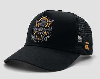 May include: Black trucker hat with a mesh back and a curved brim. The front of the hat features an embroidered design of a stylized demon face in gray, yellow, and white. The letter "a" is embroidered on the side.