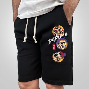 May include: Black sweatpants with a white drawstring and a graphic print of three colorful Daruma dolls with the word "Daruma" written in white. The Japanese word "元 旦" is also printed on the sweatpants.
