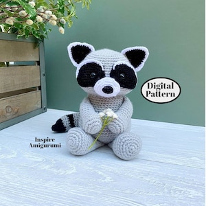 May include: A crocheted raccoon amigurumi in gray, black, and white, holding a small bouquet of white flowers. The raccoon has a black mask, ears, and tail stripes. The text "Digital Pattern" is visible. The text "Inspire Amigurumi" is also visible.