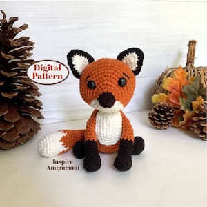 May include: A crocheted orange fox amigurumi toy with black accents, sitting on a white surface. The fox has white fur on its belly and tail tip. The toy is surrounded by fall-themed decor, including pinecones and a faux pumpkin.