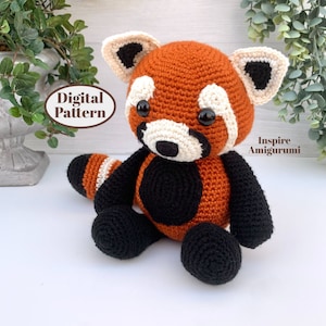 Pode incluir: Um brinquedo de amigurumi de panda vermelho feito de crochê com detalhes em preto e branco. O brinquedo está sentado em uma superfície branca. O texto "Digital Pattern" e "Inspire Amigurumi" é visível na imagem.