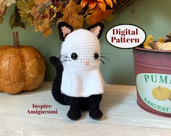 May include: A crocheted black cat wearing a white ghost costume. The cat is sitting on a wooden surface with a pumpkin and fall foliage in the background. The text "Digital Pattern" is visible in the image.