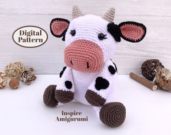 Puede incluir: Juguete de amigurumi de crochet en forma de vaca con pelaje blanco, manchas negras, pezuñas marrones y orejas y nariz rosas. El juguete está sentado sobre una superficie blanca con el texto "Inspire Amigurumi" debajo.